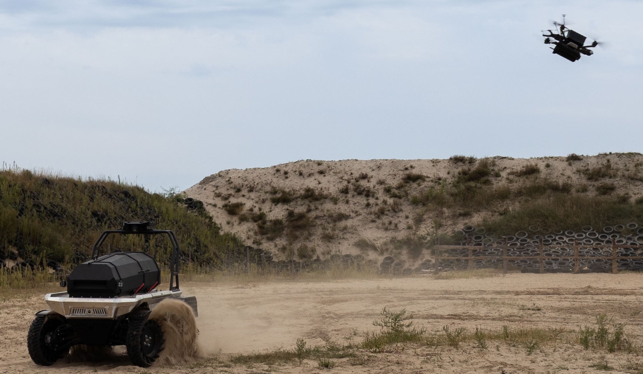 An unmanned ground vehicle kicking up sand as it drives past a wall of stacked tyres, with a reconnaissance drone overhead.