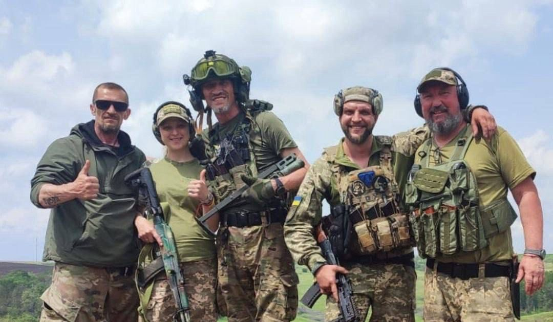 Five Ukrainian service members, one giving a thumbs up, standing together in body armour and helmets on open ground near the front line.