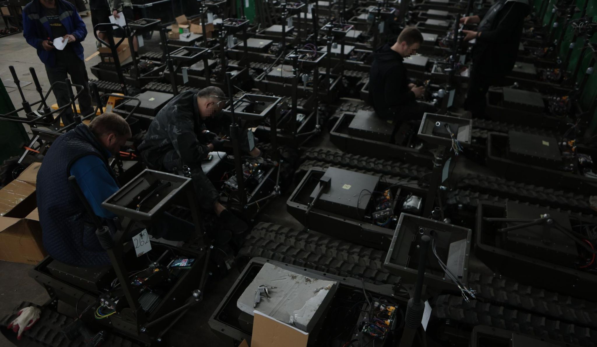 Technicians in a workshop assembling rows of tracked unmanned ground vehicles, hulls open and wiring exposed.
